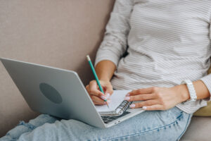 Person in a striped shirt and jeans sitting on a sofa, writing in a notebook placed on an open laptop, likely taking notes from free work from home courses.