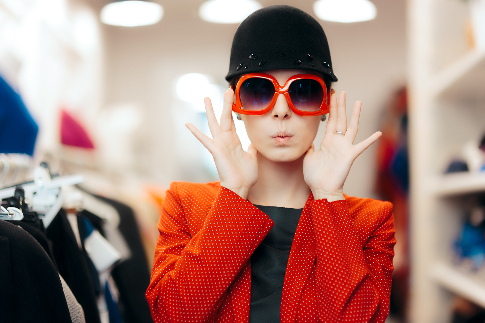 Person in a red jacket and black top, wearing large red sunglasses and a black hat, posing with hands near face, looking like a mystery shopping company's undercover agent.