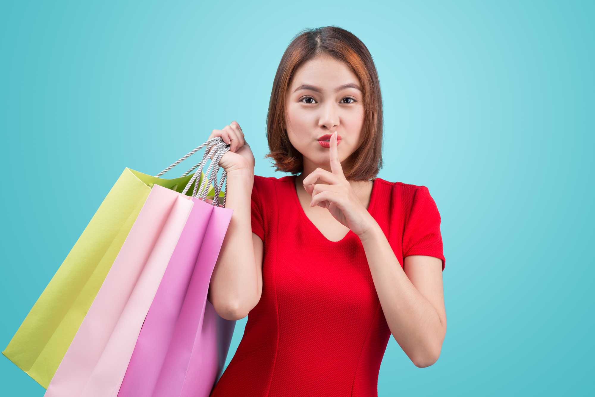 A woman in a red dress holds multiple shopping bags in one hand and makes a shushing gesture with the other hand against a solid blue background, embodying the intrigue associated with the best mystery shopping companies in the UK.