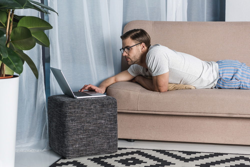 Man doing matched betting on his laptop while lying on his stomach on the sofa in pyjamas