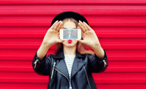 A person in a leather jacket and hat holds a smartphone, taking a selfie against a red striped background, dreaming of how they could get paid to take pictures of themselves.