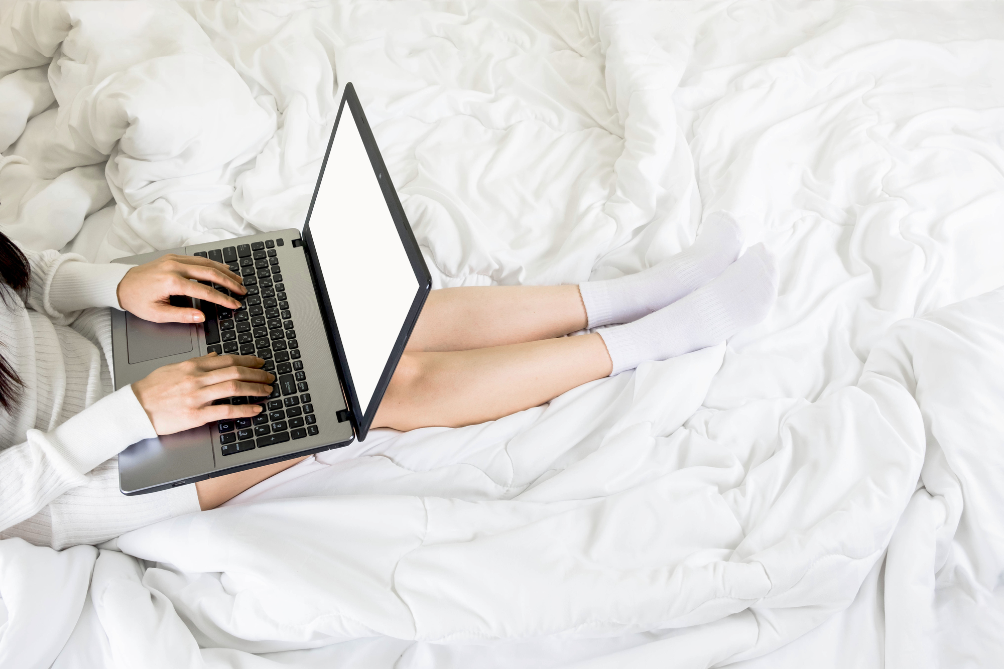 Person sitting on a bed with white sheets, typing on a laptop with a blank screen, wearing white socks and a light-colored outfit. The scene subtly hints at researching "how to make money without a job.