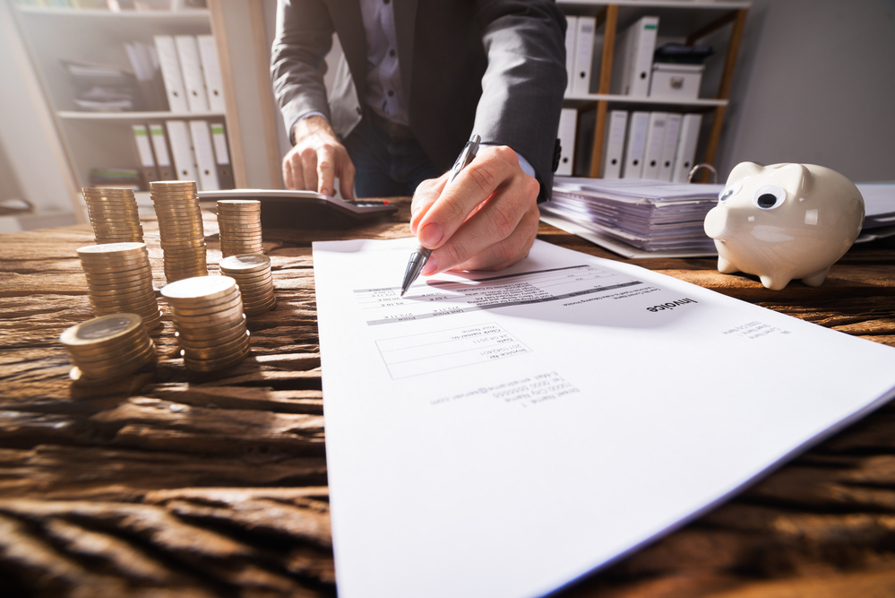 Close-up Of A Businessperson's Hand Signing Document With Stacked Golden Coins On Wooden Desk