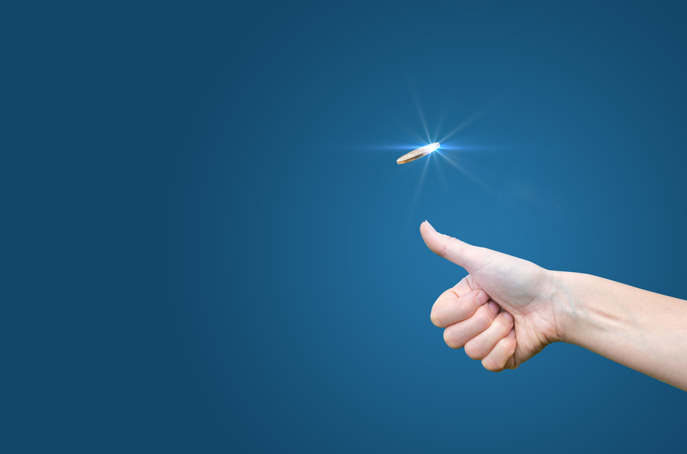 A hand flipping a coin against a blue background, a simple form of gambling used to demonstrate matched betting