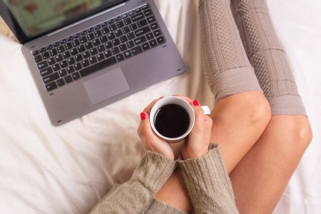 A person in knitted socks and a sweater sits on a bed holding a cup of coffee, with a laptop nearby, perhaps researching how to make money without a job.