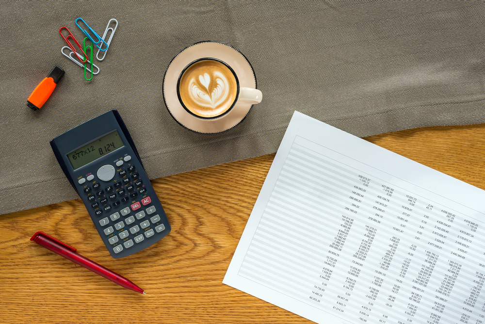 flat lay of budgeting spreadsheet, calculator, coffee and paper clips on a wooden desk. It's important to budget carefully if you do matched betting full time.