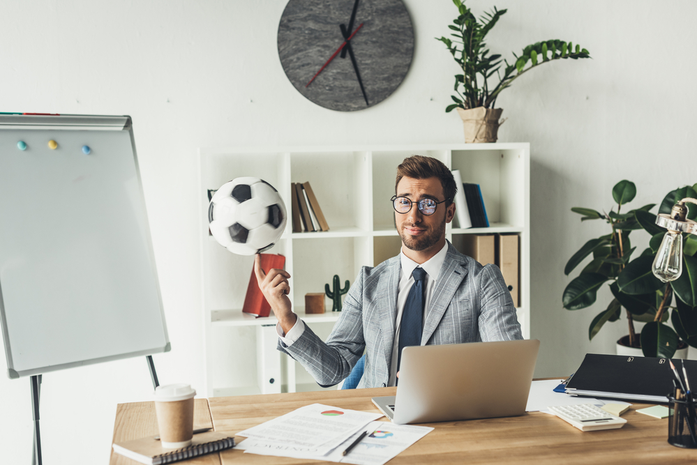 A businessman spinning a soccer ball on his finger while reading matched betting tips at his desk.