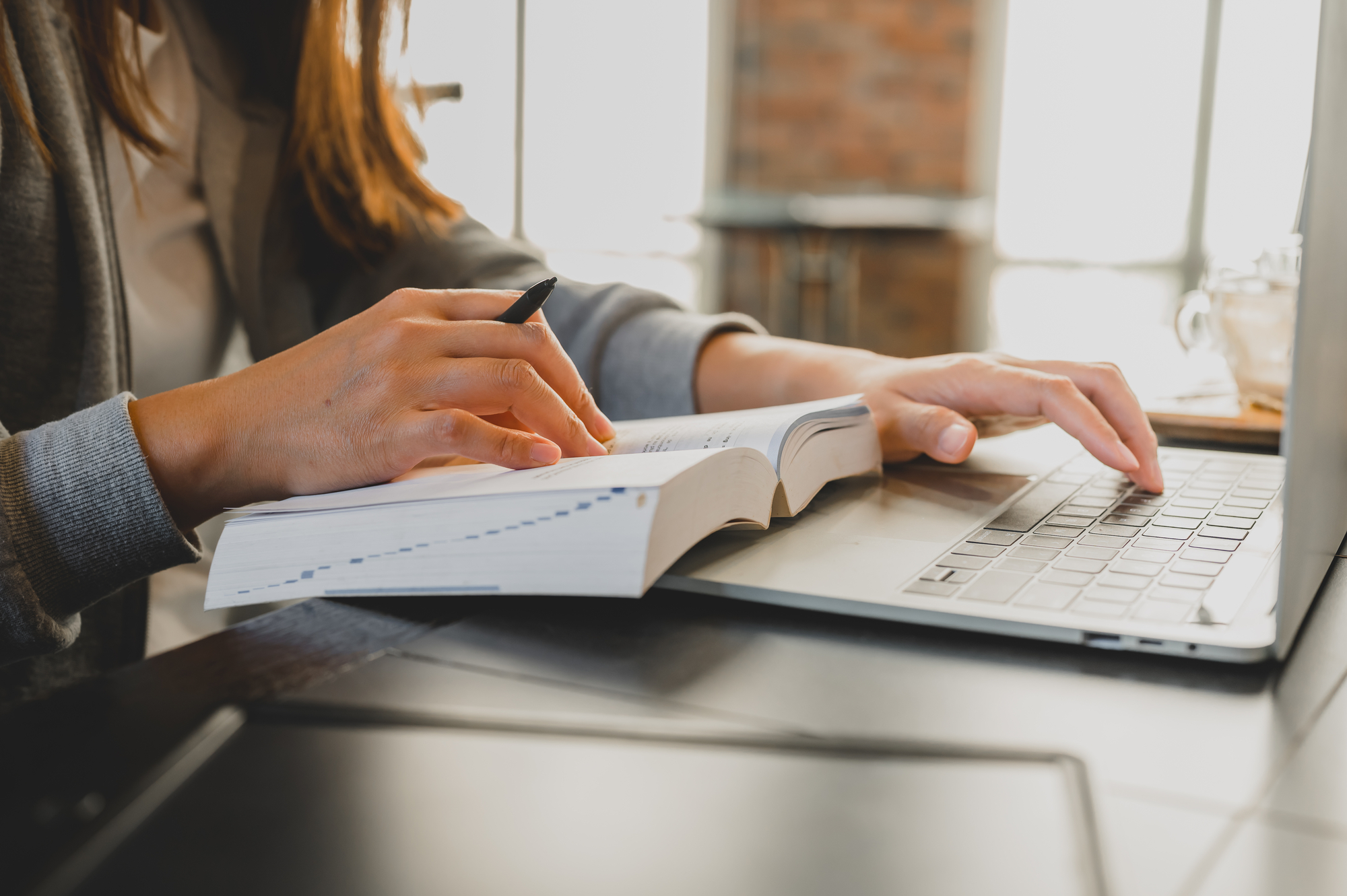 A woman multitasking between referring to a large dictionary and working on her laptop, exploring online jobs for students.