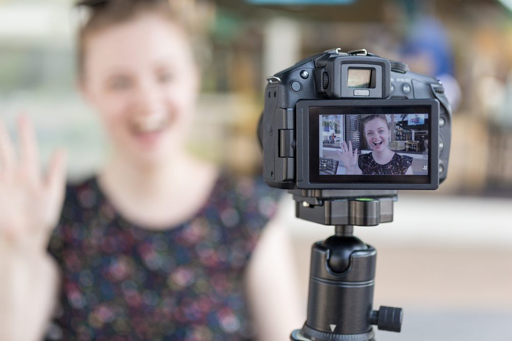 A woman in the UK is waving to the camera while holding a video camera for her youtube side hustle.