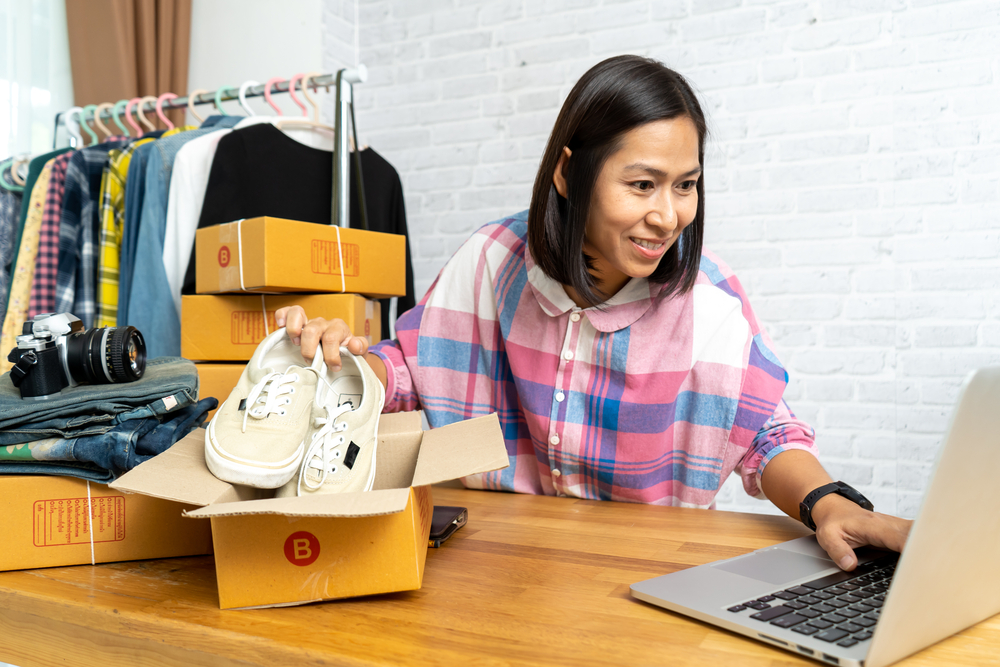 A woman in the UK is browsing a laptop near a stack of clothes which she is packing into boxes for her reselling side hustle.