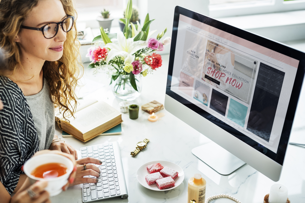 a woman at a side hustle in the UK, sitting at a desk with a cup of tea and design ideas on the computer screen.