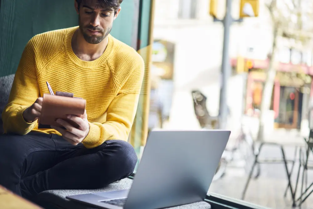 a man in yellow jumper making notes on a notepad, sitting by a laptop