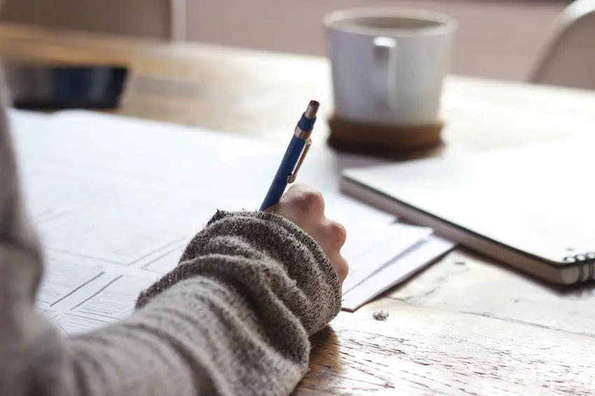 A hand holding a pen writing on some papers with coffee on desk. earn extra money marking exams