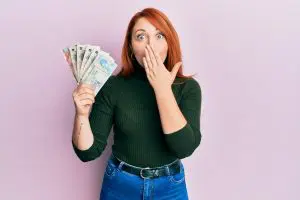 a woman holding a handful of Uk pound sterling notes and covering her mouth in surprise