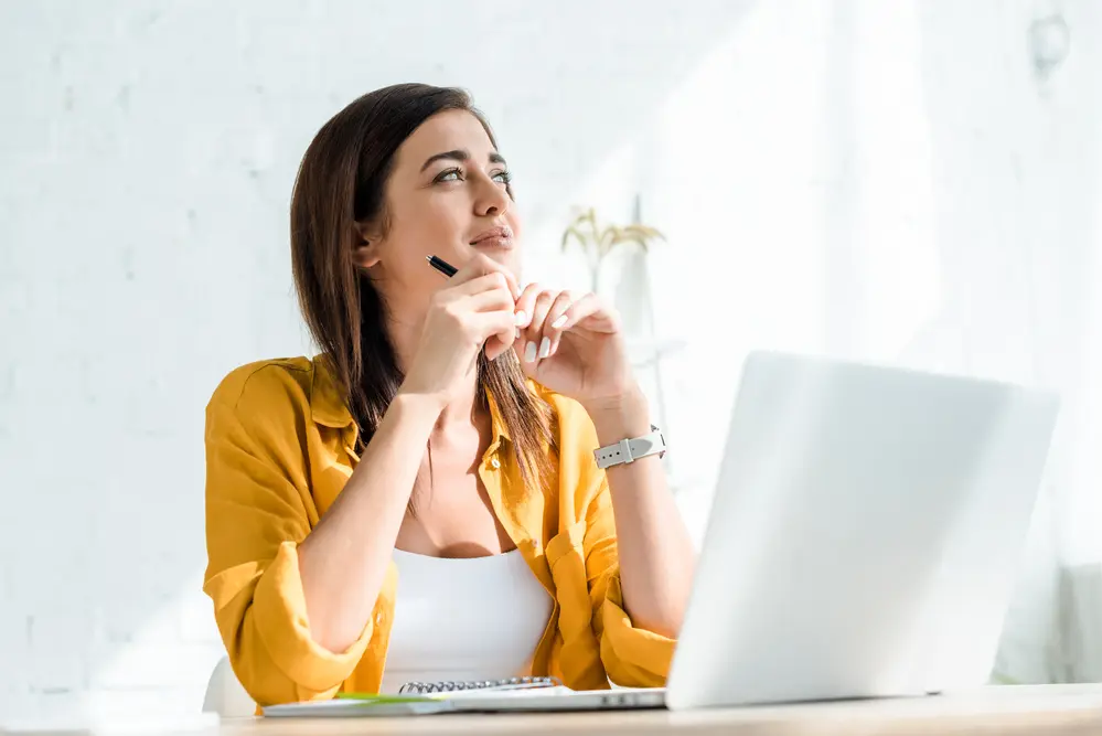 a freelance woman sitting at her desk with a laptop in front of her and a pen in her hand, thinking