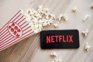 A smartphone displaying the Netflix logo is placed next to a popcorn container with spilled popcorn on a wooden surface, reminiscent of those moments when you're figuring out how to get paid to watch Netflix all day.