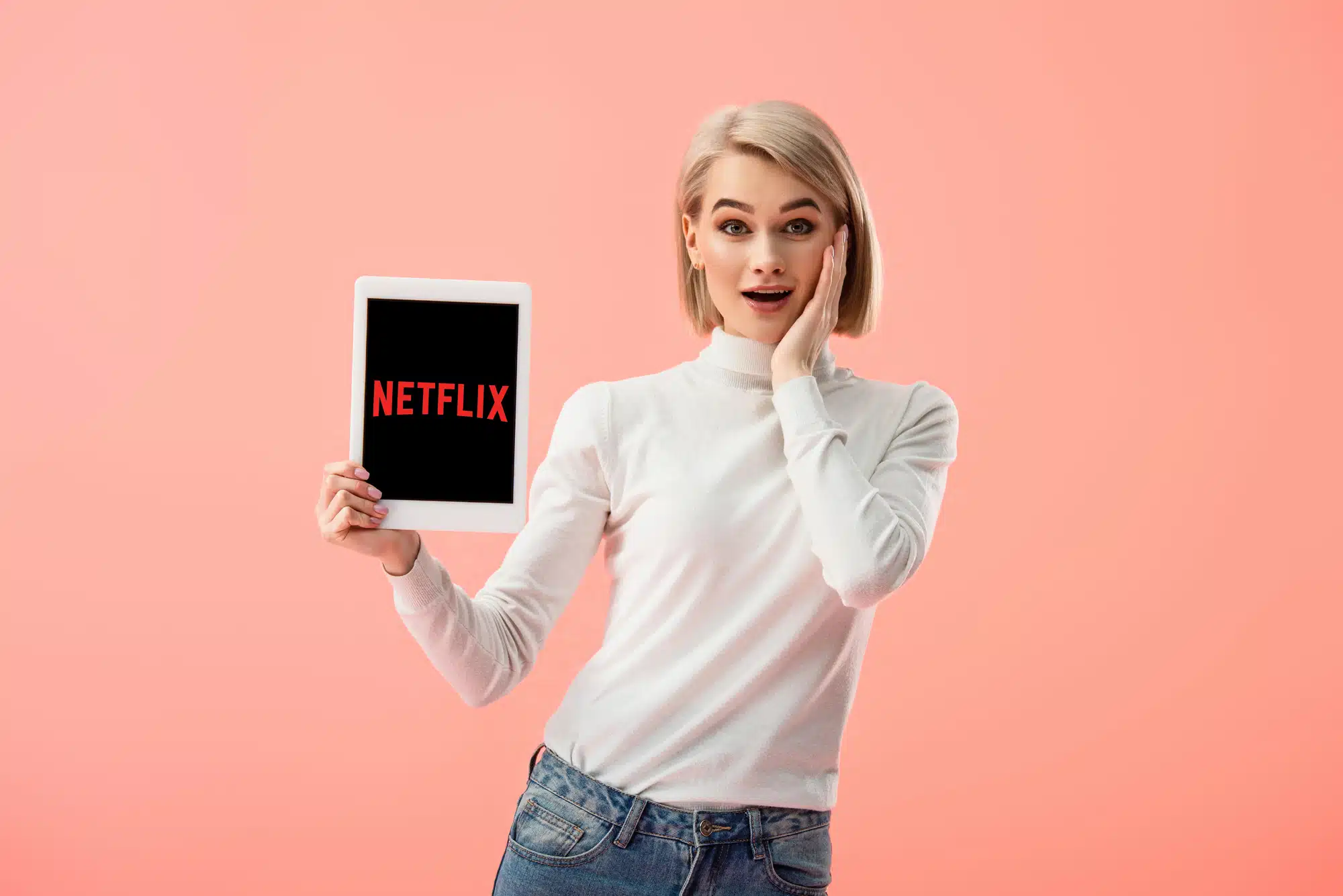 A woman with blonde hair, wearing a white turtleneck and jeans, holds a tablet displaying the Netflix logo against a pink background, with a surprised expression as if discovering how to get paid to watch Netflix.