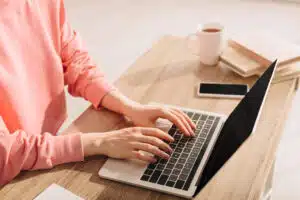 Person in a pink sweater typing on a laptop at a wooden desk with a smartphone, a cup of coffee, and a few books nearby, exploring online jobs that require no experience.